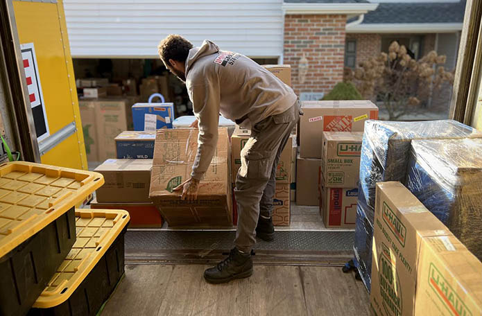 A mover in a branded hoodie bends to lift a cardboard box from the back of a moving truck. The truck is surrounded by tightly packed labeled boxes.