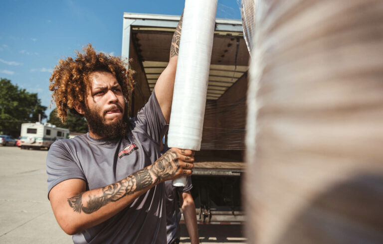 A mover with tattoos and curly hair stretches packing film around furniture, standing beside an open moving truck under a clear sky.