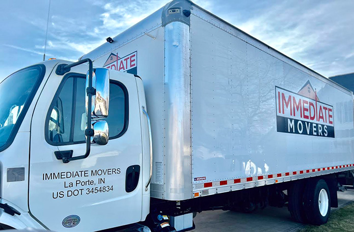 A white Immediate Movers & Storage box truck is parked in the driveway of a suburban home surrounded by trees with autumn leaves on the ground.